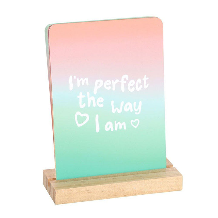 Affirmation card on wooden stand, pink-teal gradient: "I'm perfect the way I am"