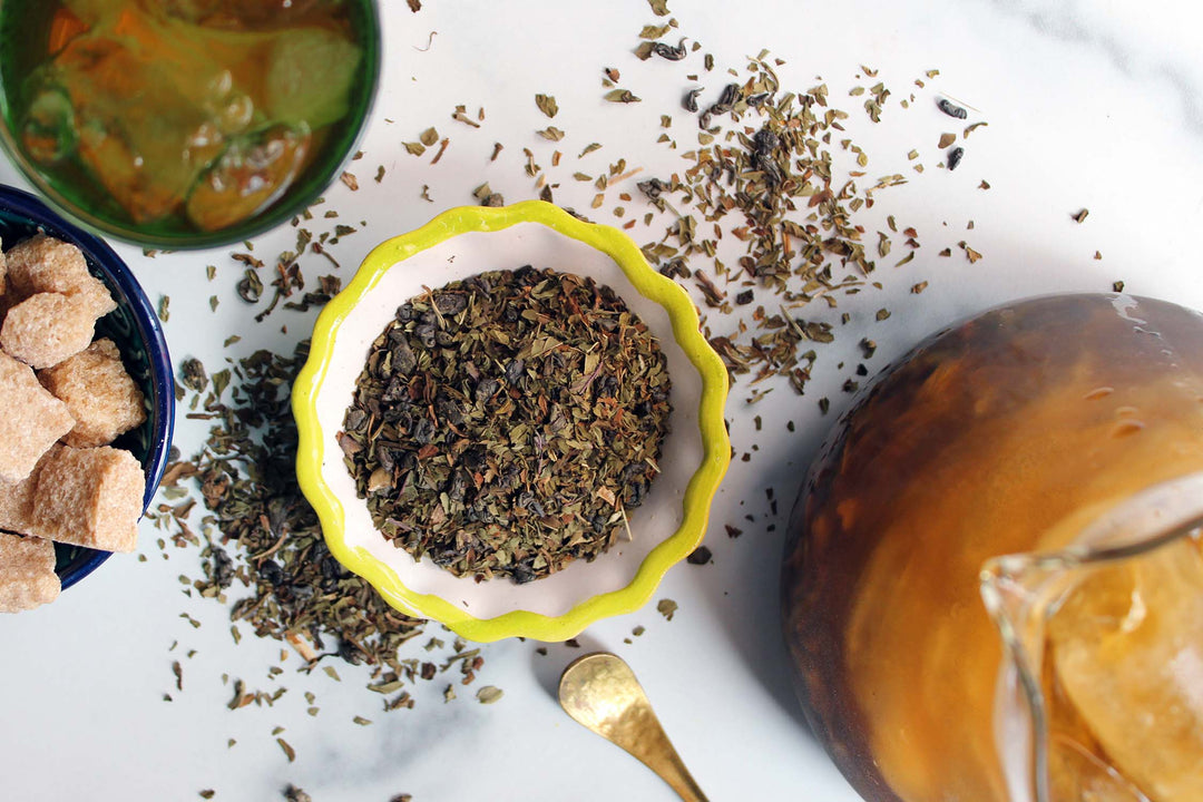 Bowl of dark tea leaves with scattered mint shreds and a spoon on a white surface.