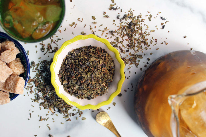 Bowl of dark tea leaves with scattered mint shreds and a spoon on a white surface.