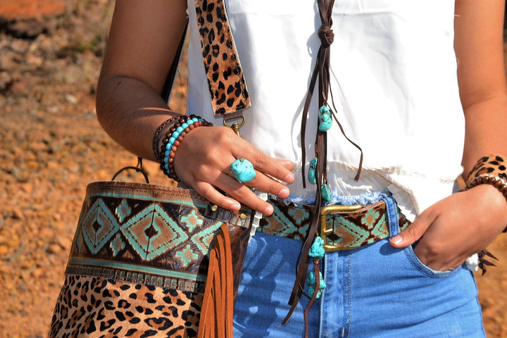 Turquoise cuff ring on hand near patterned bags and denim jeans