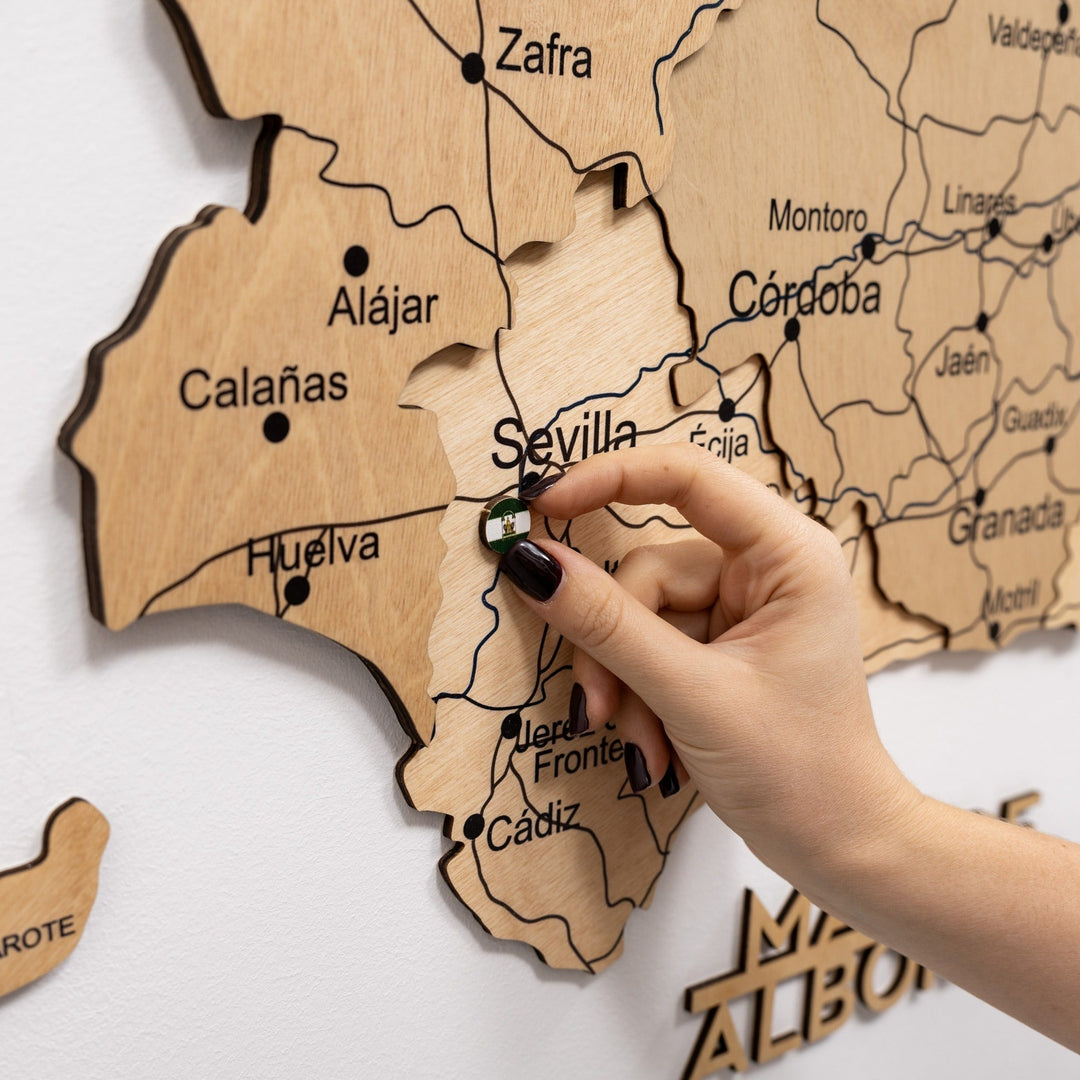 Hand places a pin on gold wooden Spain map near Sevilla; street lines and labels visible