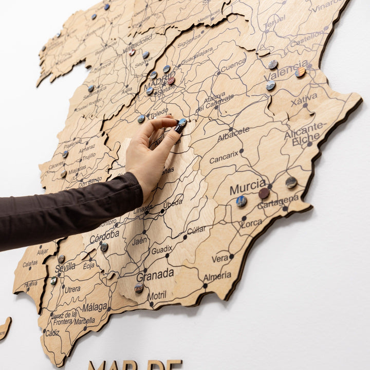 Hand placing a pin on gold wooden Spain map; city names and road lines visible
