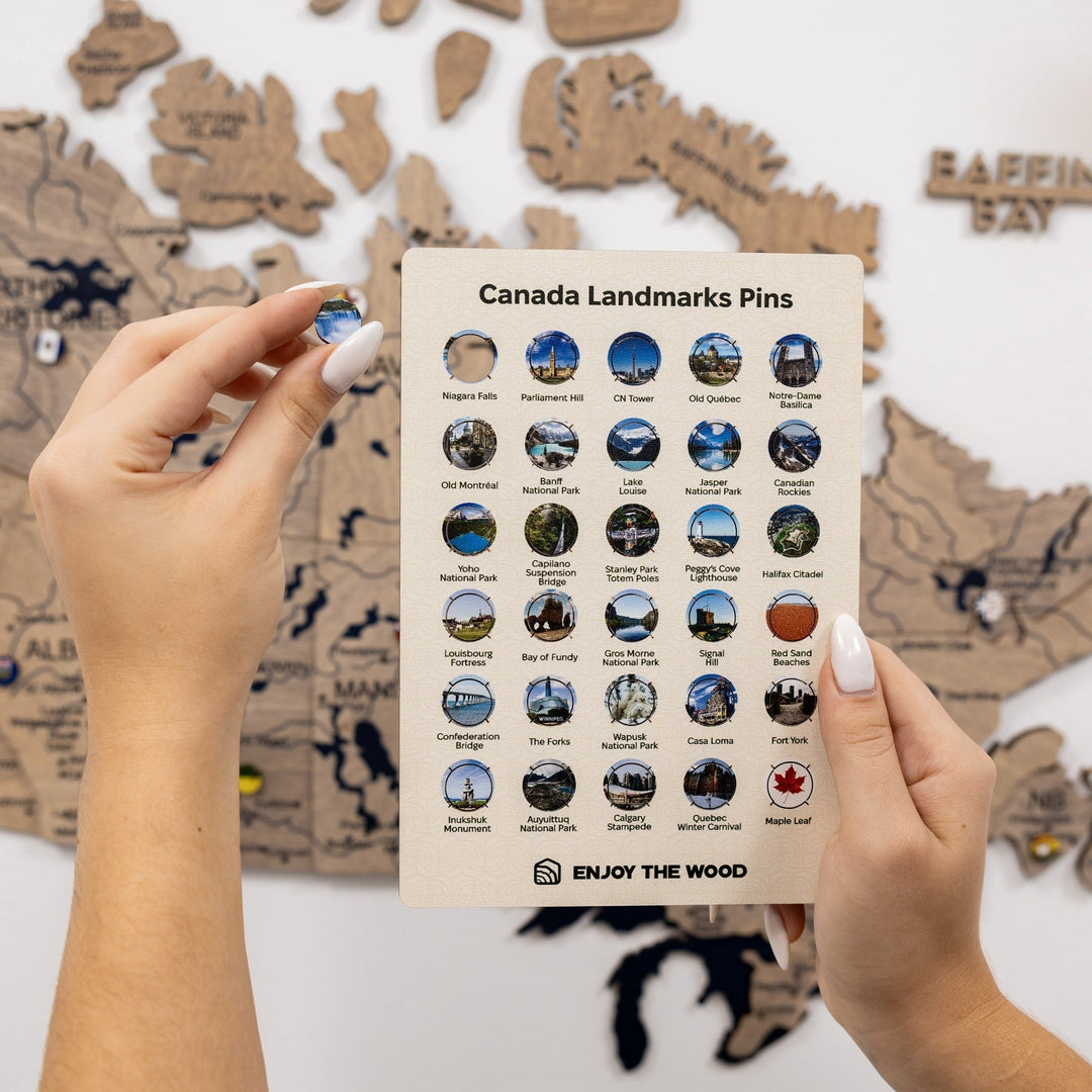 Hands holding a pin display card labeled "Canada Landmarks Pins" over the wooden map.