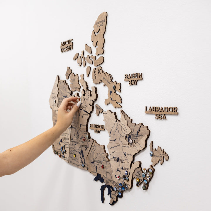 Wooden Canada wall map with raised regions and pins while a person places a pin.