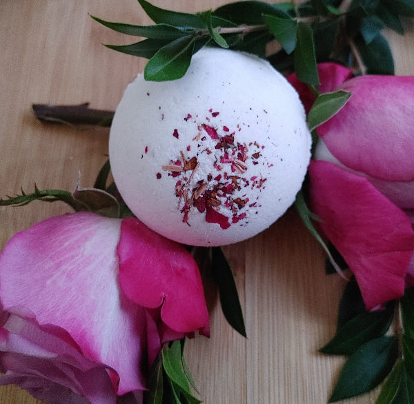 Round bath bomb with red-pink petals, placed among pink flowers and green leaves