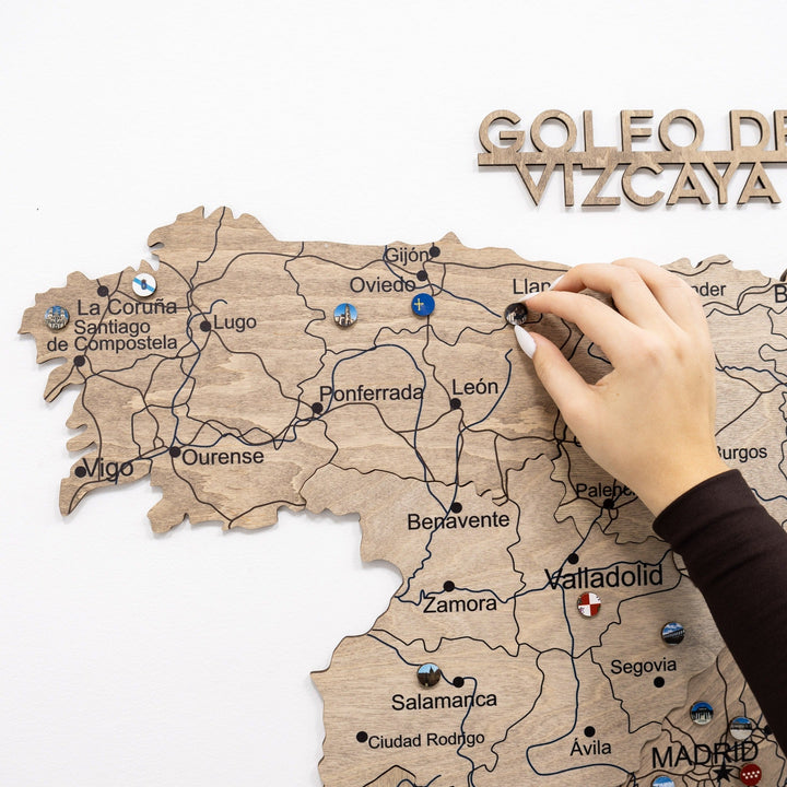 Spain landmark enamel pins on wooden map board, hand attaching round pin near labels