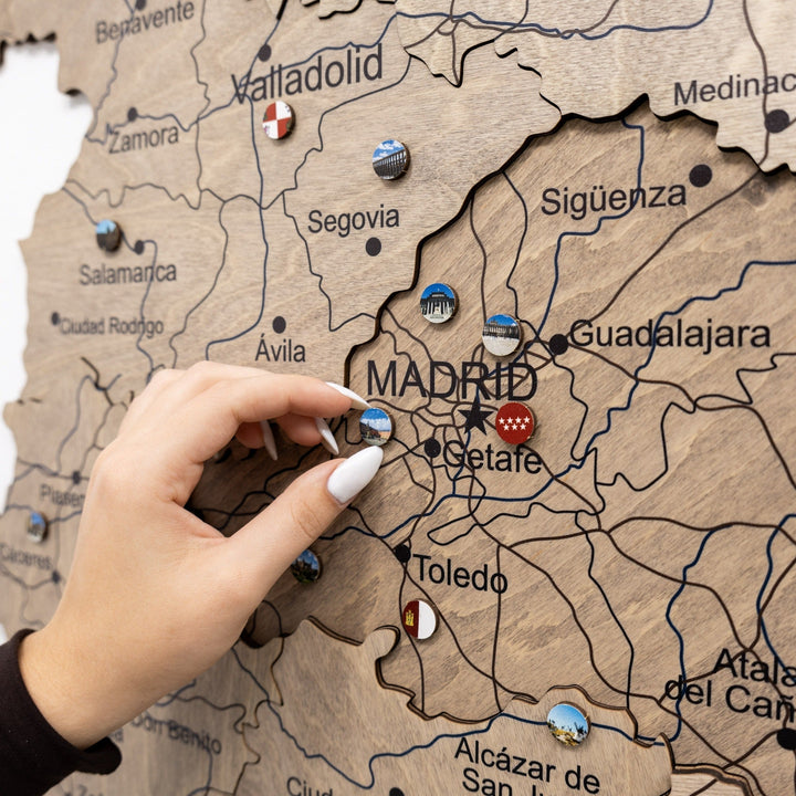 Round Spain landmark enamel pins attached to wooden map, hands pressing into place