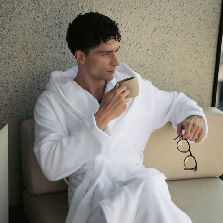 White plush hooded bathrobe with tie belt, worn by a seated man; holding glasses.